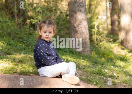 Piccola bella ragazza si siede su una superficie di pietra in un parco su uno sfondo naturale sfocato verde Foto Stock