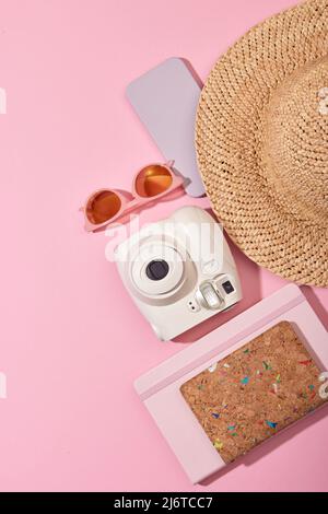 Da sopra di fotocamera istantanea con occhiali da sole smartphone e cappello di paglia posto vicino al notebook su sfondo rosa Foto Stock