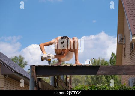 Un falegname lavora con un cacciavite elettrico giallo, ruota una vite in una trave di legno della tavola sul tetto Foto Stock