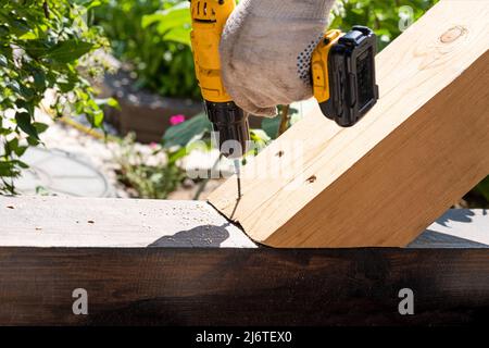 Un falegname lavora con un cacciavite elettrico giallo, ruota una vite in un asse di legno trave. Foto Stock