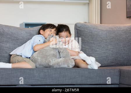 Una bambina e un ragazzino giocano felicemente con una bambola di elefante ripiena sul divano del soggiorno - foto di scorta Foto Stock