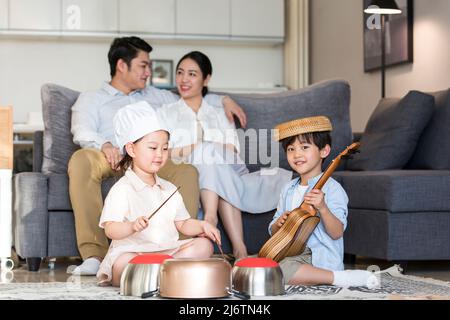 Un ragazzo e una ragazza si siedono sul tappeto giocando mischievously su ukulele e utensili da cucina, mentre i genitori guardano in ammirazione felice - foto di scorta Foto Stock