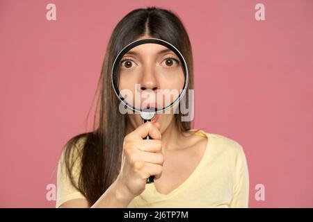 Primo piano ritratto di giovane donna stupita guardando attraverso una lente di ingrandimento su sfondo rosa Foto Stock