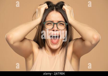 Ritratto closeup stressato donna frustrata con occhiali urlando isolato su sfondo beige. Reazione negativa dell'espressione facciale dell'emozione umana atti Foto Stock