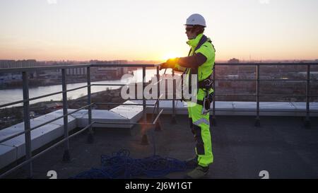 Il climber industriale sul tetto srotea la corda di sicurezza e appende la matassa sulle spalle. Uomo in verde chiaro tuta e casco si prepara per il lavoro al tramonto Foto Stock