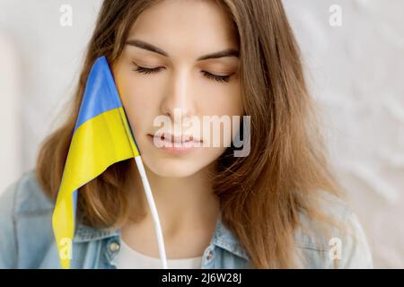 La ragazza seria indipendente tiene la bandiera Ucraina e gli occhi vicini. Patriot del suo paese. Prega per l'Ucraina. Lotta per la libertà. Stop guerra. Blu giallo. Foto Stock