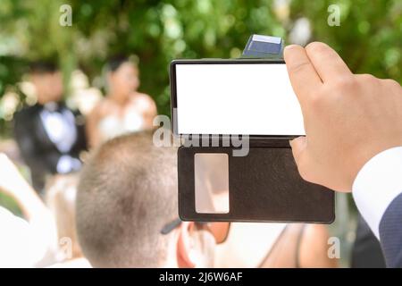 Schermo con spazio copia di un telefono cellulare tenuto dalla mano di un ospite che sta registrando il matrimonio di una coppia di nozze all'aperto in estate. Foto Stock