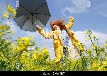 Donna con una camicia blu e un impermeabile giallo che salta nel campo della colza gialla e si diverte in una giornata estiva soleggiata Foto Stock