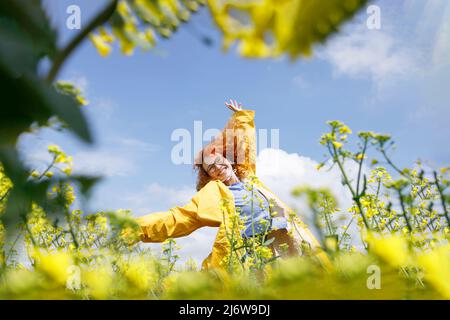 Una donna in un impermeabile giallo che salta nel campo dei fiori gialli e si diverte in una giornata di sole Foto Stock