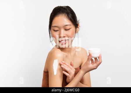 Donna asiatica sorridente che applica crema corpo sulla spalla Foto Stock