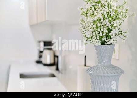 Vista ravvicinata dei piccoli fiori bianchi della gypsophila in elegante vaso grigio sul piano di lavoro della cucina, senza gente. Camera moderna con sfondo interno Foto Stock