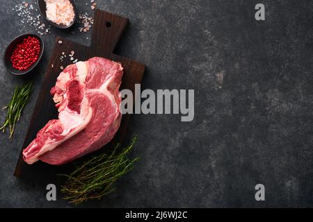 Carne di manzo cruda. Pezzo grezzo di carne su osso per arrosti o zuppa con sale, pepe, timo e rosmarino su fondo di cemento nero. Entrecote. Cupolino grezzo Foto Stock