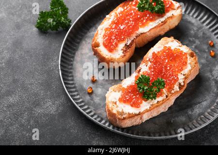 Due dandwiches con caviale rosso. Caviale rosso salmone in ciotola e sandwich server su vecchio piatto di ferro su sfondo vecchio tavolo nero. Vista dall'alto. Spazio di copia. Foto Stock