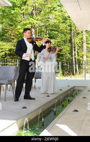 Duo di violinisti. Un uomo in un cappotto di coda e una donna in un abito da sera giocare il violino vicino alla fontana. Foto Stock