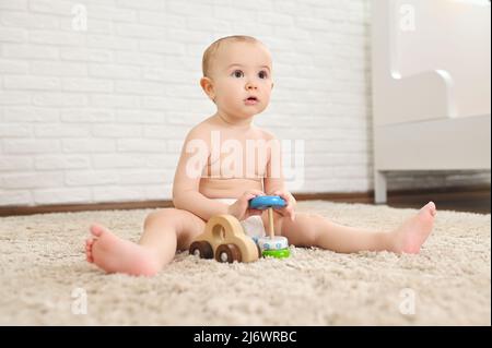 il bambino cute siede su un tappeto in un pannolino usa e getta gioca con i giocattoli contro una parete di mattone bianca. Foto Stock