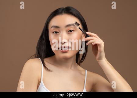 Affascinante signora asiatica che applica il mascara del sopracciglio, usando il trucco decorativo e posando alla macchina fotografica sopra lo sfondo marrone dello studio Foto Stock