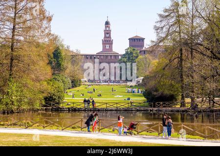 Milanese e turisti si godono il Parco Sempione di Milano, dominato dalle torri del Castello Sforzesco. Foto Stock