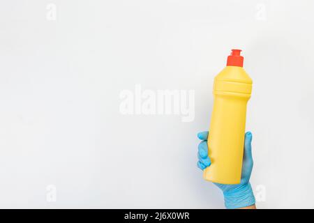 La mano di una donna della pulizia in un guanto di gomma blu tiene una bottiglia di detergente su uno sfondo grigio. Detersivo per varie superfici nel angolo cottura Foto Stock