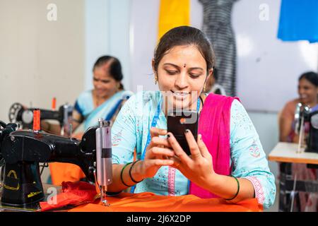 Donna su misura utilizzando il telefono cellulare di fronte alla macchina da cucire a indumenti - concetto di rilassamento, tecnologia e controllo online disegni. Foto Stock