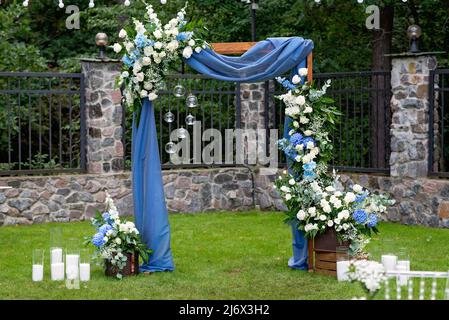 Luogo per una cerimonia nuziale all'aperto. Rustico arco in legno decorato con tessuto blu, fiori e candele. Messa a fuoco selettiva soft. Foto Stock