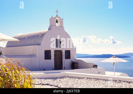 Oia, Santorini, Isole Greche, Grecia - Chiesa greco-ortodossa con campanile con ombrelloni in scintillante luce del sole che si affaccia sul mare Foto Stock