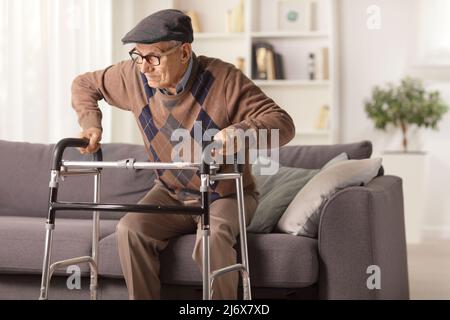 Uomo anziano su un divano cercando di camminare con un camminatore a casa Foto Stock