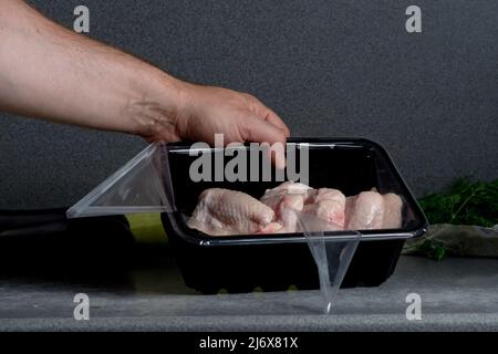 Una mano maschile tiene le ali di pollo crudo in un involucro di plastica sul tavolo da cucina. Preparazione per la cottura. Foto Stock