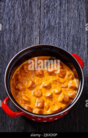 nakkikastike, salsicce finlandesi, amalgami in salsa di pomodoro cremosa in pentola rossa su tavola di legno scuro, vista verticale dall'alto, posa piatta, spazio libero, primo piano Foto Stock