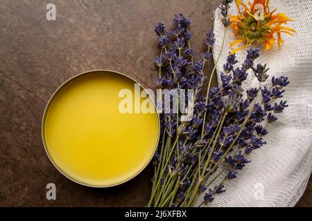 Immagine closeup presa da sopra di una scatola di calendula fai da te fatta in casa e salsa di lavanda accanto alle erbe essiccate e cheesecloth su sfondo marrone mattonelle. Foto Stock