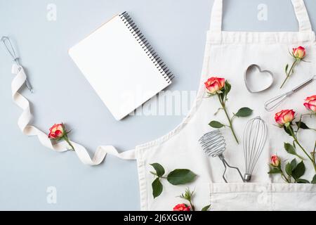 Libro di ricette vuoto, accessori da forno con grembiule con fiori rose su sfondo grigio, piatto, vista dall'alto. Ricette di cottura festive Foto Stock