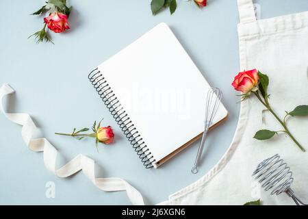 Libro di ricette vuoto, accessori da forno con grembiule con fiori rose su sfondo grigio, piatto, vista dall'alto. Ricette di cottura festive Foto Stock