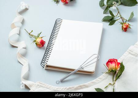 Libro di ricette vuoto, accessori da forno con grembiule con fiori rose su sfondo grigio, piatto, vista dall'alto. Ricette di cottura festive Foto Stock