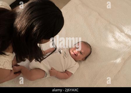 La giovane madre latina bacia i piedi del bambino e li annidano sul letto di casa Foto Stock