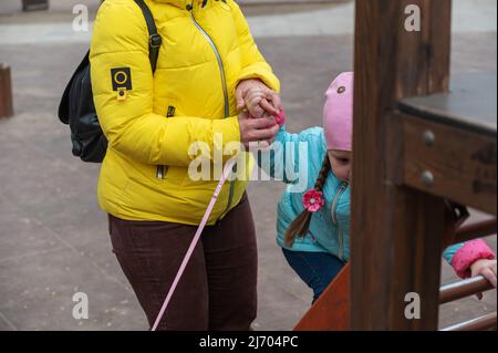 La mamma aiuta la figlia a salire lo scivolo dei bambini. Una donna cresciuta con una giacca gialla sta tenendo un guinzaglio rosa per il suo cane. Una ragazza con pigtail lunghi. Foto Stock