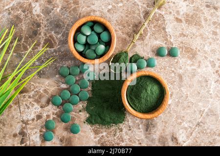 Polvere di spirulina verde e compresse in ciotole di legno su sfondo marmoreo marrone. Un superfood preferito per vegetariani e atleti. Vista dall'alto Foto Stock