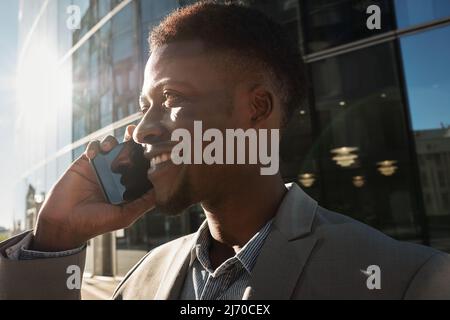 Primo piano ritratto del giovane manager afroamericano che parla su smartphone e sorride allegramente sul sfondo del business center Foto Stock