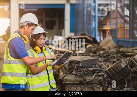 Giovane uomo asiatico e donna staff lavoratore che lavora in cantiere sporco blocco motore rottami. Team di ingegneri che utilizzano il computer tablet insieme. Foto Stock