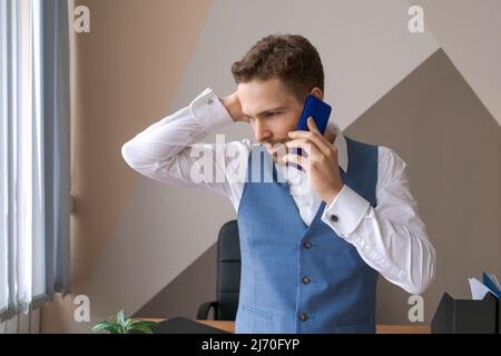 Piacevole bell'uomo in camicia e gilet che parla al telefono, ordinando taxi o consegna di cibo in ufficio. Sorridente giovane uomo d'affari caucasico che tiene una chiamata con i clienti partner. Foto Stock