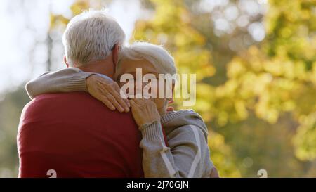 Abbraccia tra due anziani caucasici. Uomo in maglione rosso visto dalla schiena, la moglie dai capelli grigi abbraccia il collo. Foto di alta qualità Foto Stock