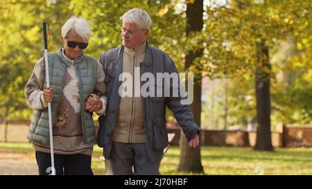 Vita con disabilità. Una coppia anziana che in autunno ha fatto una passeggiata nel loro parco preferito. Signora cieca disabile con canna bianca e occhiali da sole supportati dal suo amato marito. Foto di alta qualità Foto Stock