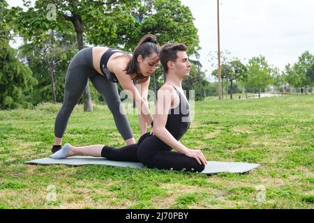 Allenatore personale femminile che aiuta il giovane uomo in un parco con posa di piccione yoga. Foto Stock