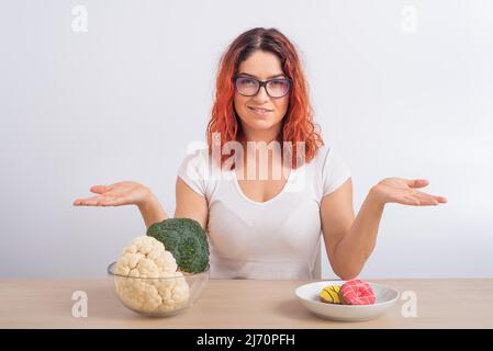 La donna caucasica preferisce cibo sano. Ragazza rossa sceglie tra broccoli e ciambelle su sfondo bianco. Foto Stock