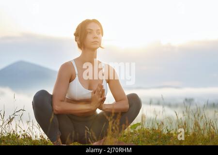 Vista panoramica frontale di giovane donna seduta in lotos posa in collina. Bella donna che fa yoga, guardando avanti con le mani di preghiera. Concetto di armonia con la natura. Foto Stock