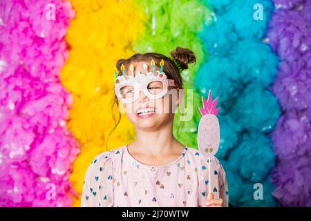 Ragazza carina che festeggia il compleanno, indossando occhiali di carta da festa, ridendo e divertendosi. Sfondo luminoso color arcobaleno. Felice evento. Foto Stock