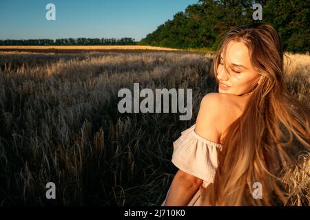 Connettiti con la natura, rallenta, sia presente, entra nei tuoi sensi. Tranquilla da sola giovane donna in abito a piedi sul campo di grano al tramonto giorno d'estate Foto Stock