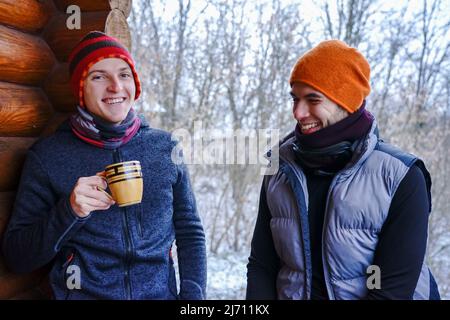 Due ragazzi chiacchierano dopo lo sci. Si trovano sul balcone di una casa di legno in abiti invernali, bere tè e rilassarsi. Portatore di due ragazzi in una stazione sciistica in Foto Stock