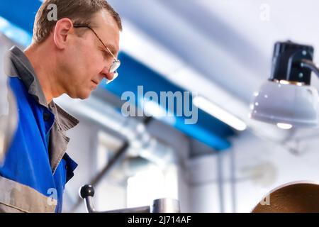 Ritratto di Turner con vetri 50-55 anni al lavoro. La torcente in metallo anziano lavora in officina su una macchina dietro il lavoro. Scena reale. Lavoratore reale. Foto Stock