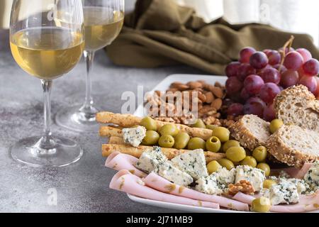 Due bicchieri di vino bianco e la piastra con stuzzichini vari. Formaggio blu, olive, baguette a fette, grissini, prosciutto, uva e i dadi. Vino set snack backg Foto Stock
