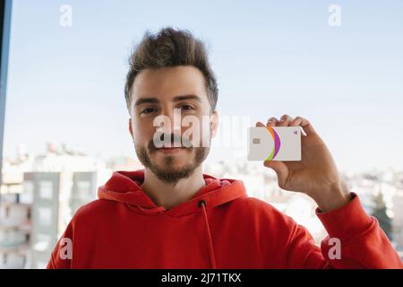 Giovane uomo bello con cappuccio rosso che mostra una carta di credito o di debito. Concetto di shopping online o di digital banking. Fuoco selettivo sull'uomo. Foto Stock