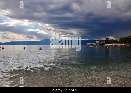 Bella spiaggia croata baia con barche nel mare di smaragd. Città di Opatija e montagna Ucka sullo sfondo. Tempesta estiva che arriva su patina e mare Adriatico. Foto Stock